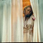 Eine Frau im Seidenkleid steht am Fenster, schaut durch den Vorhang. Der Hintergrund ist orange beleuchtet.