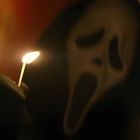 Ein schwarzer behandschuhter Hand hält ein brennendes Streichholz vor einer gruseligen Maske.