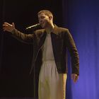 Ein Mann singt in ein Mikrofon, trägt eine gestreifte Jacke und helles Shirt, vor einem dunklen Hintergrund.