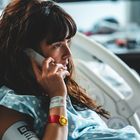 Text: „Wichtiger Alarm Warnarmband auf Blutdruck Armplatzierung“. Eine Frau im Krankenhausbett telefoniert, schaut besorgt.