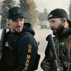 Zwei uniformierte Männer mit Ausrüstung. Ein Mann trägt ein Kleidungsstück mit "U.S. MARSHAL". Sie sind in ernster Unterhaltung.