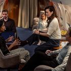 Mehrere Personen sitzen in einer luxuriösen Flugzeugkabine und diskutieren angeregt. Ein Mann hält ein Glas Wasser.