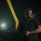 Keine erkennbaren Texte. Ein Mann steht nachts draußen, hält etwas. Im Hintergrund Baustellenfahrzeug und Licht.