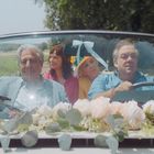 Vier Personen sitzen in einem Cabrio, das mit Blumen geschmückt ist. Sie fahren auf einer Landstraße.