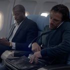 Zwei Männer sitzen in einem Flugzeug. Einer schaut auf sein Handy, der andere telefoniert und hält eine Tasche.