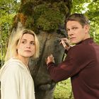 Ein Mann schnitzt mit einem Messer Schrift in einen Baum, daneben steht eine Frau. Sie sind von Blättern umgeben.