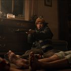 Ein Mann zielt sitzend mit einem Gewehr. Links steht eine Lampe auf einer Kommode. Zwei Personen liegen am Boden.