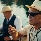 Zwei Männer in Strohhüten und Sonnenbrillen sitzen am Wasser. Der Vordergrundmann hält eine Pfeife.