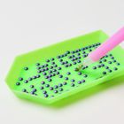 Kein Text. Ein grünes Tablett mit lilafarbenen Steinen und ein rosa Plastikstift, der einen Stein aufsammelt.