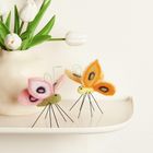 Zwei Filzschmetterlinge in Pink und Orange stehen auf Drahtbeinen neben einer Vase mit weißen Tulpen.