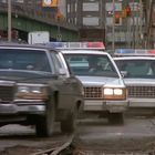 Zwei Polizeiautos mit Blinklichtern verfolgen ein schwarzes Auto auf einer Straße. Im Hintergrund sind Verkehrsampeln.