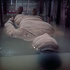 In einem flachen Wasserbecken liegt eine Person mit bandagiertem Körper und Kopf, umgeben von einem metallischen Gestell.
