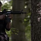 Ein Mann in schwarzer Kleidung zielt mit einem Gewehr im Wald. Die Beleuchtung ist schattig durch die Bäume.