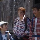 Drei Personen, ein Kind und zwei Erwachsene, in Freizeitkleidung mit Rucksäcken, stehen vor einer Holzhütte.