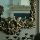 Keine sichtbaren Texte. Ein Spiegelbild zeigt eine Puppe mit großen Augen in einem kunstvoll verzierten, goldenen Rahmen.