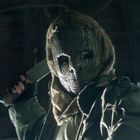 Ein maskierter Mensch mit einer groben, strukturhaften Maske hält ein Messer, umgeben von dunkler, unscharfer Umgebung.