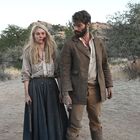 Eine Frau mit blutverschmiertem Gesicht und ein Mann im Westernstil stehen in einer trockenen, buschigen Landschaft.