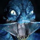 Ein blaues, reptilienartiges Wesen mit großen Augen und einem weit geöffneten Maul zeigt scharfe Zähne.