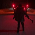 Person steht nachts auf schneebedeckter Straße vor einem Fahrzeug, hält eine Axt, Scheinwerfer leuchten rot.