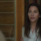 Eine Frau mit langen, dunklen Haaren steht in einem Raum mit Holzwänden und schaut ernst aus dem Bild.