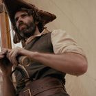 Zwei Männer auf einem Segelschiff bei starkem Wind, einer hält ein Fernrohr, mit Segeln und Masten im Hintergrund.