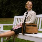 Eine Frau in einem beigen Mantel sitzt lächelnd auf einer weißen Bank im Park, hält eine Tasse, mit Füßen ausgestreckt.