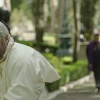 Ein älterer Mann in weißer religiöser Kleidung mit goldenem Kreuz läuft. Im Hintergrund folgen weitere Personen auf einem Weg.