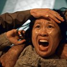 Eine Frau in Panik, Mund weit geöffnet, umgeben von mehreren Händen, die sie festhalten. Licht und Schatten verstärken die Dramatik.