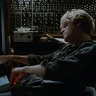 Links bunte Retro-Animation auf einem Bildschirm. Mann mit hellem Haar vor großem Synthesizer, Getränkedose neben ihm.