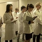 Sechs Personen in weißen Laborkitteln stehen in einem Flur, halten Klemmbretter und schreiben Notizen.