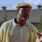 Ein Mann im Freien trägt einen beigen Hut, ein helles Poloshirt mit einem kleinen Krokodil-Logo und eine gelbe Jacke.