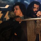 Zwei Männer, einer hält den anderen mit einem Arm im Schwitzkasten, der andere mit einer Stange. Ernsthafte Stimmung.