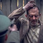 Es gibt keinen Text. Ein Mann hebt die Hände, während eine Person mit Mütze eine Pistole auf ihn richtet. Er wirkt besorgt.