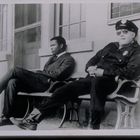 Zwei Männer auf einer Bank. Links ein Mann im Anzug, rechts ein Polizist mit Uniform und Sonnenbrille. Im Hintergrund "Waiting".