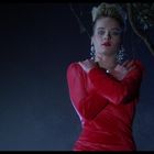 Kein sichtbarer Text. Eine Frau in rotem Kleid, Arme gekreuzt, steht vor dunklem Hintergrund mit Baum und dunklem Himmel.