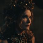 Keine sichtbaren Texte. Eine Frau in seitlicher Ansicht trägt eine kunstvolle Krone. Sie hat langes, welliges Haar und einen ernsten Ausdruck.