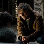 Ich kann den Text im Bild nicht erkennen. Ein Mann mit lockigem Haar lächelt und kniet vor einer Person in einem dunklen Raum.