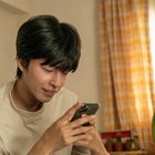 Ein Mann in einem beigen T-Shirt schaut lächelnd auf sein Handy. Im Hintergrund sind Pflanzen und gelb karierte Vorhänge.