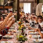 Viele Menschen sitzen an einem langen Tisch und heben Gläser zum Anstoßen. Der Tisch ist festlich gedeckt mit Blumen.