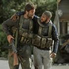 Zwei Männer in Tarnwesten stützen sich, einer hält ein Gewehr. Sie wirken erschöpft, im Hintergrund grüne Vegetation.