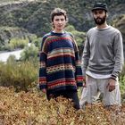 Keine Texte sichtbar. Ein Mann und eine Frau stehen in einer hügeligen Landschaft, umgeben von Büschen und einem Fluss im Hintergrund.