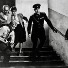 Vier Personen rennen eine Treppe hinunter. Einer trägt eine Uniform, die anderen wirken besorgt und eilig.