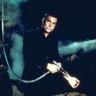 Es gibt keinen sichtbaren Text. Ein Mann steht in einer dunklen Umgebung und hält einen Metallstab. Rauch im Hintergrund.