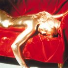 Eine goldene Gestalt liegt auf rotem Stoff, ausgestreckt auf einem Sofa. Ein eindrucksvolles Bild mit glitzernder Oberfläche.