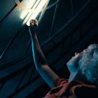 Ein blonder Mensch in orangefarbener Kleidung streckt sich nach einer Lichtquelle in einer dunklen Umgebung mit futuristischen Elementen.