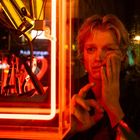 Neonschriftzüge, eine Person drückt ihr Gesicht gegen eine Glasscheibe, umgeben von warmem Neonlicht.