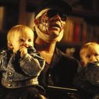 Eine Person mit verbundenem Gesicht hält zwei Babys in Jeansjacken; im Hintergrund sind Bücher zu sehen.
