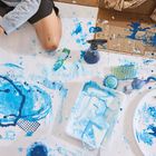 Kinderhände malen mit blauem und weißem Farbchaos auf einem großen Papier. Verschiedene Malwerkzeuge sind verstreut.