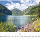 "Frühling am Fjord; Monatskalender für Mai. Blühende Wiesen vor einem Fjord mit steilen, grünen Bergen unter blauem Himmel."