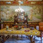 Wände mit kunstvollen Gemälden, großer hölzerner Esstisch mit bunter Tischdecke, dekorativer Kronleuchter.